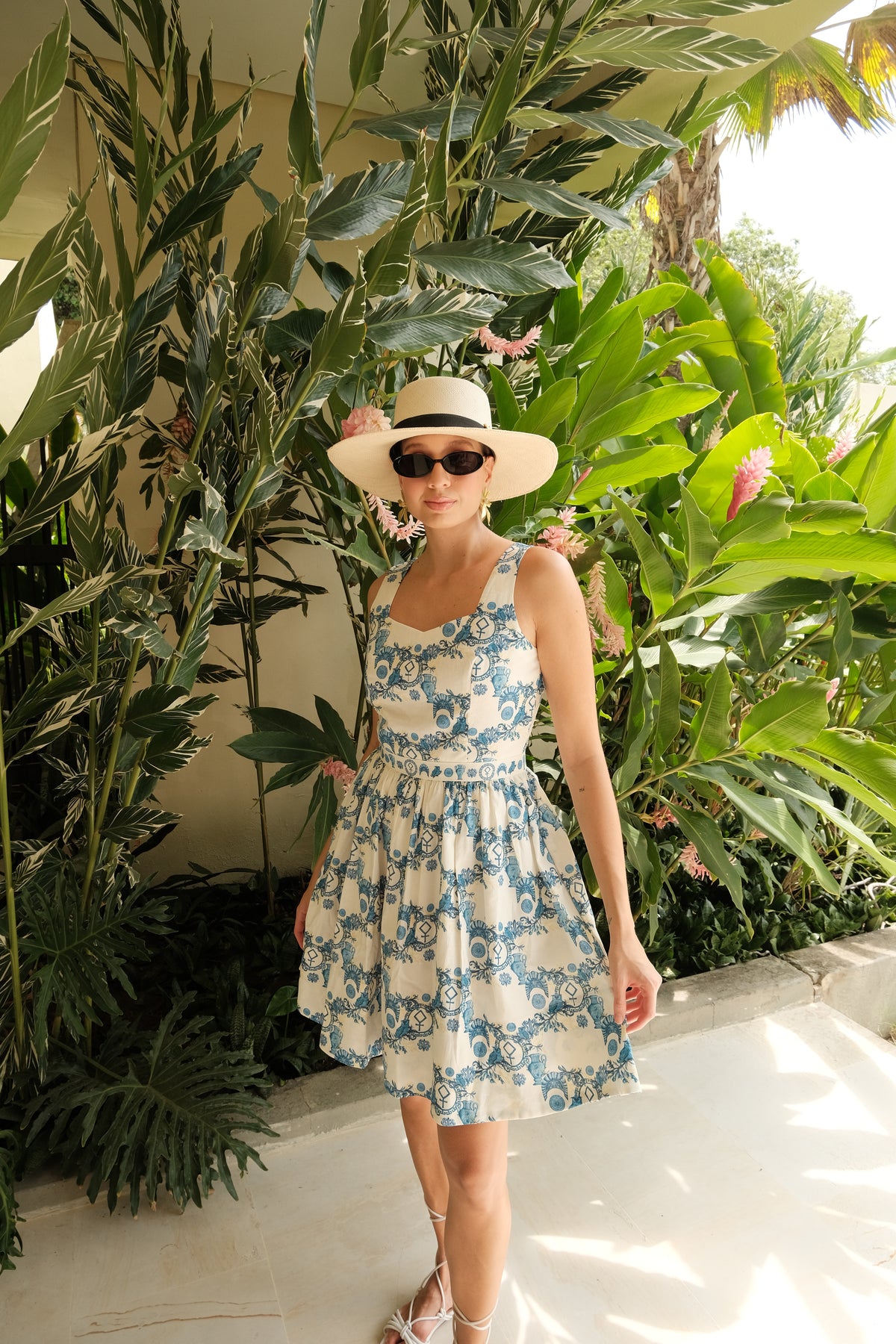 A woman wearing Athena's Triumph blue and white hemp and cotton sleeveless dress, a wide-brimmed straw hat, and sunglasses, posing outdoors in front of lush tropical green plants during a day time party. 