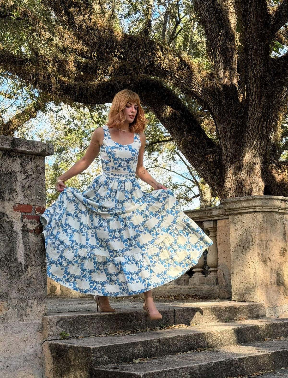 A woman wearing a blue and white floral print sleeveless dress stands on stone steps outdoors under a large tree, holding out the sides of the dress to show its full flowy silhouette.