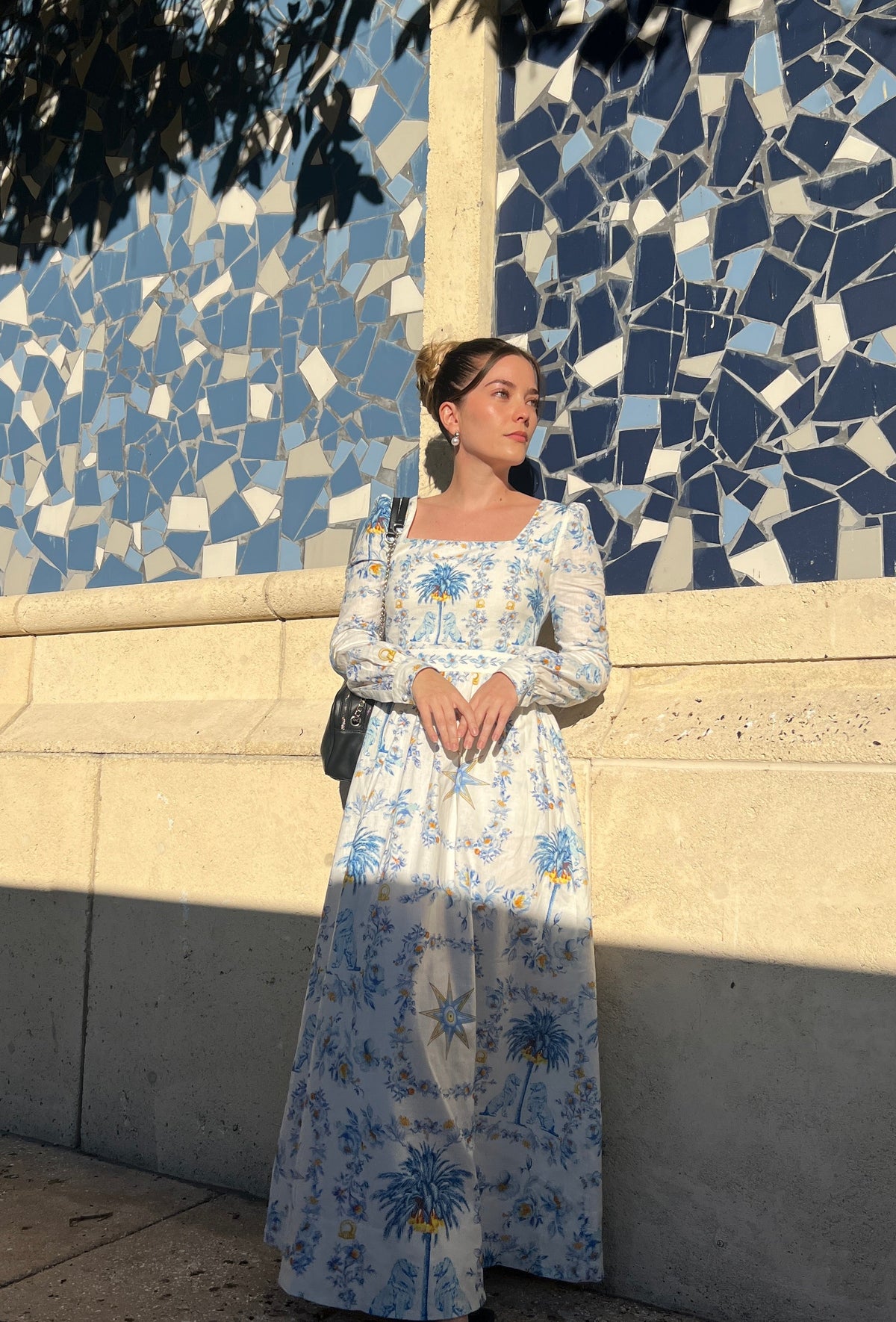 Woman wearing Ishtar linen and cotton maxi dress against a blue and white mosaic wall in warm late afternoon light. The dress has long voluminous sleeves gathered into buttoned cuffs, a square neckline framing the shoulders, a fitted bodice with a defined waistband, and a full-length straight skirt. White ground with celestial blue and golden saffron print featuring the eight-pointed Star of Ishtar, date palms, and Sumerian motifs. Hair worn in a low bun, pearl drop earring visible, small dark crossbody bag