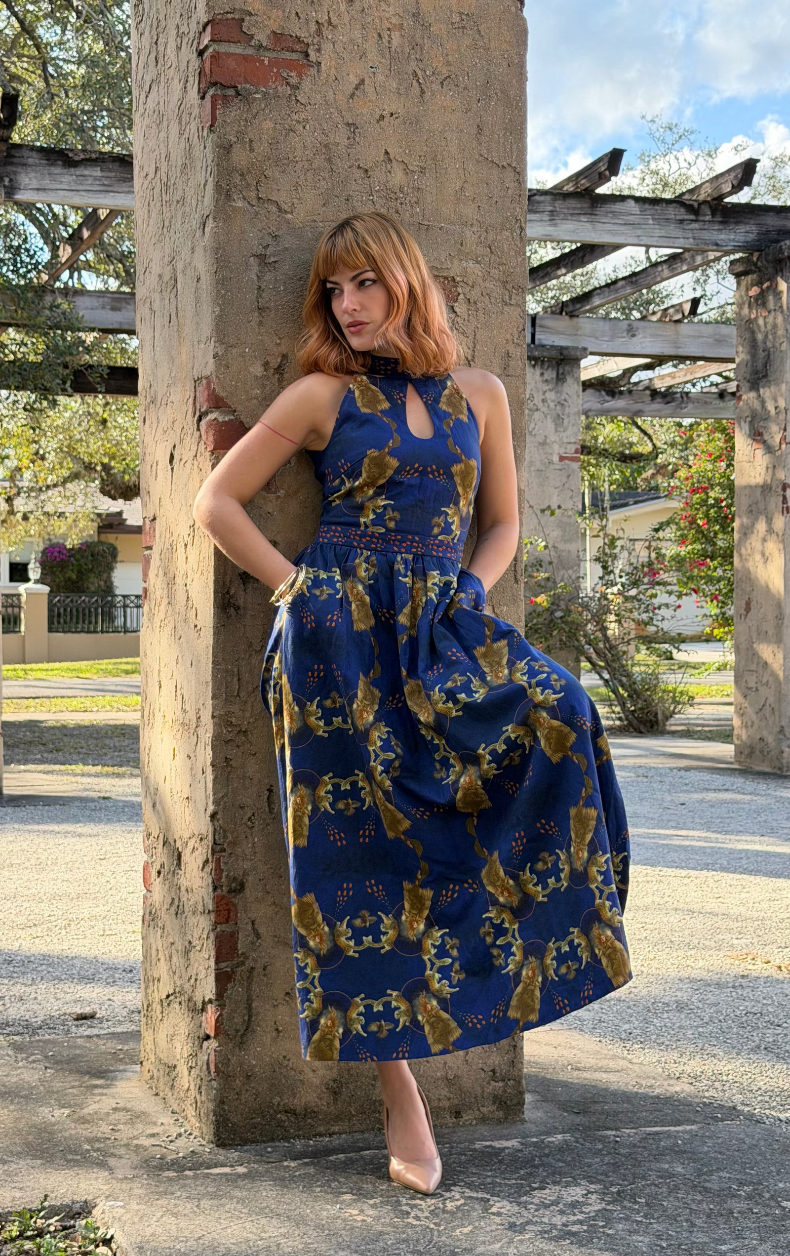 Diva Thea model wearing a sleeveless blue maxi dress with a gold pattern leans against a rustic concrete pillar in an outdoor garden setting in Miami.