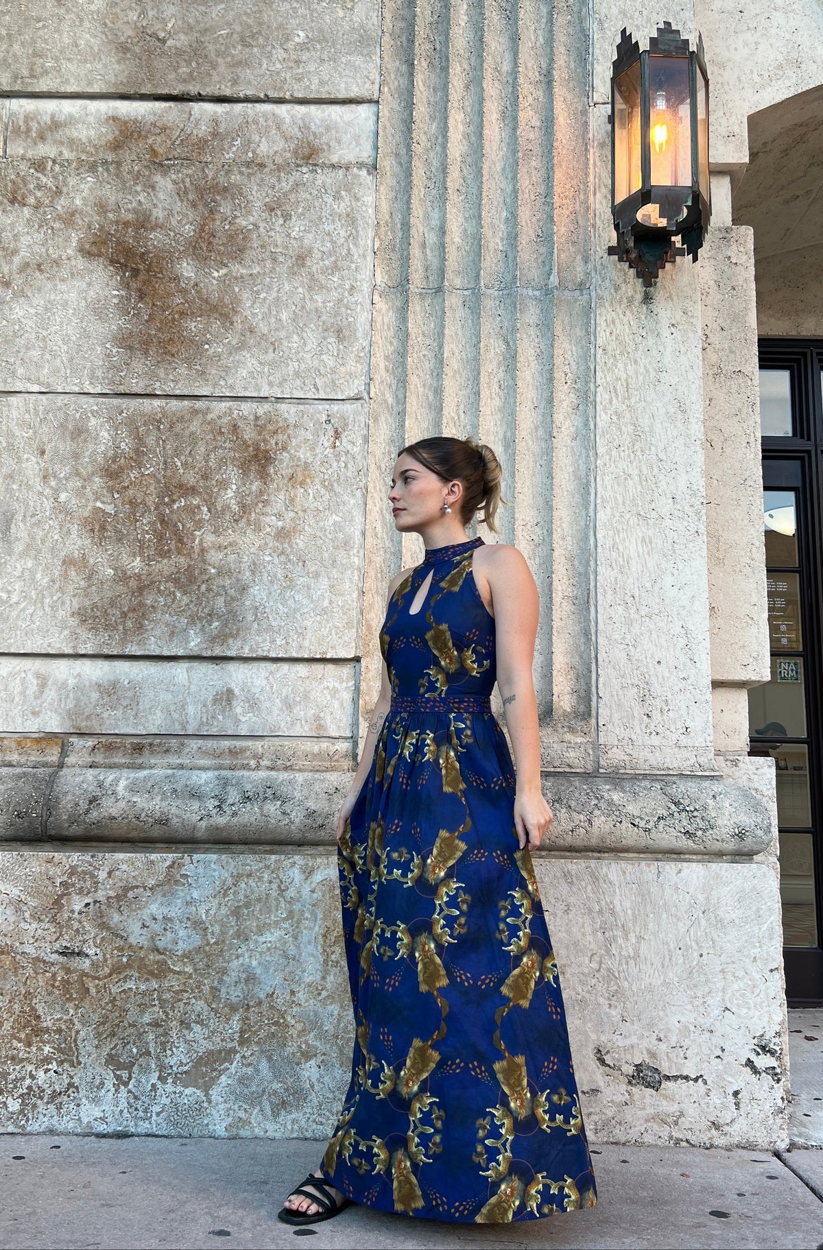 A woman wearing a sleeveless navy blue halter maxi dress with a gold pattern featuring Norse goddess Freya pattern, standing in front of a stone building column. Location: Coral Gables, Florida