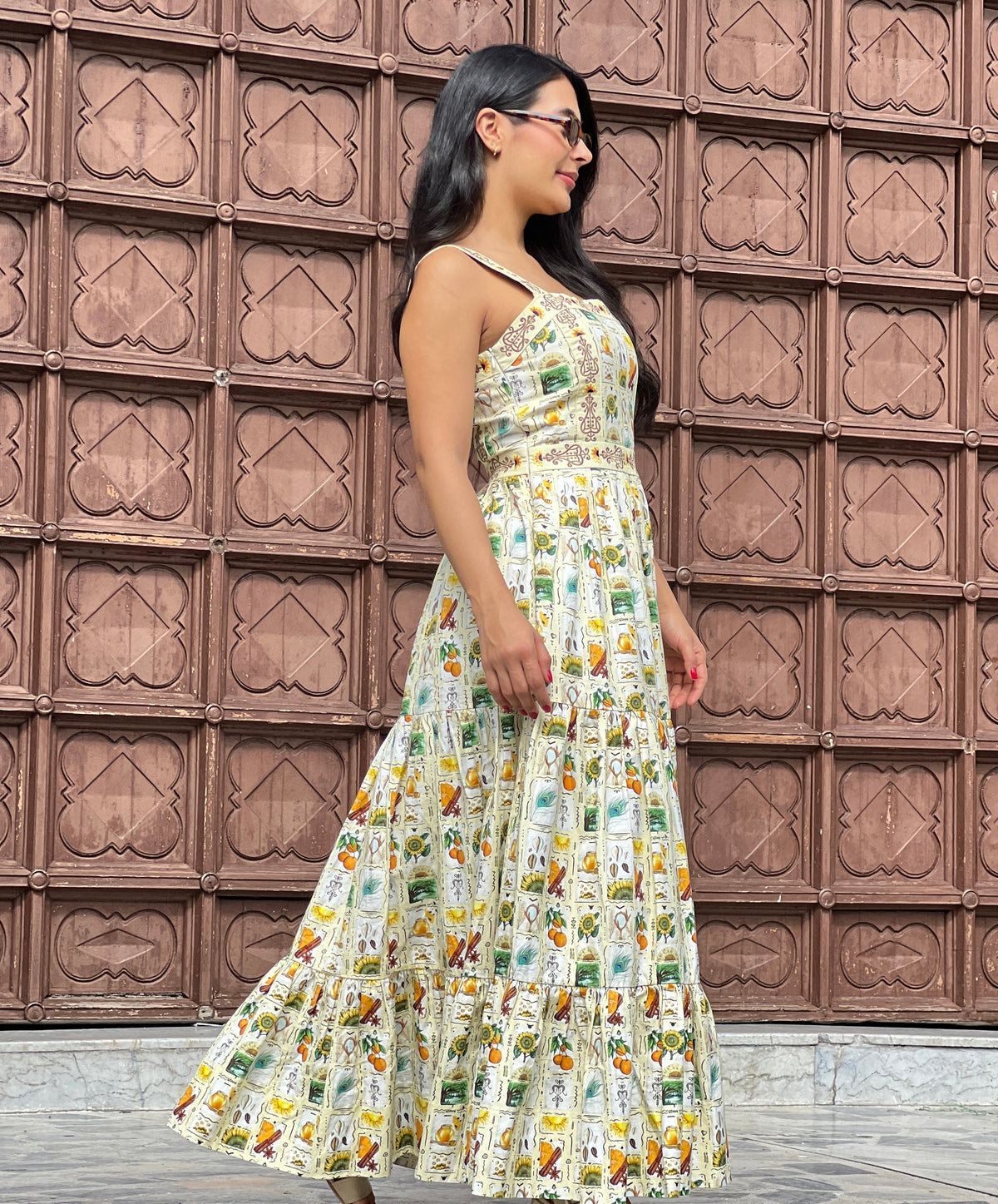 Woman shown in side profile wearing the Oshun's Celebration maxi dress. Sleeveless cotton maxi with thin shoulder straps, shaped neckline, fitted bodice with embroidered waistband, and three-tiered gathered skirt. Cream ground printed with original Yoruba-inspired vignettes depicting sunflowers, cowrie shells, honey jars, oranges, cinnamon, candles, mirrors, peacock feathers, and rivers. Background shows a carved wooden colonial door, suggesting warm-weather travel or cultural event wear.