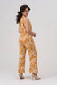 Women’s wide-leg halter jumpsuit in natural linen, featuring Diva Thea’s Amber Radiance print in gold, amber, and deep ochre. Adjustable halter neckline, fitted natural waist without elastic, tiered wide-leg silhouette, and side pockets. Suitable for weddings, cultural events, warm-weather dinners, and other occasion dressing.